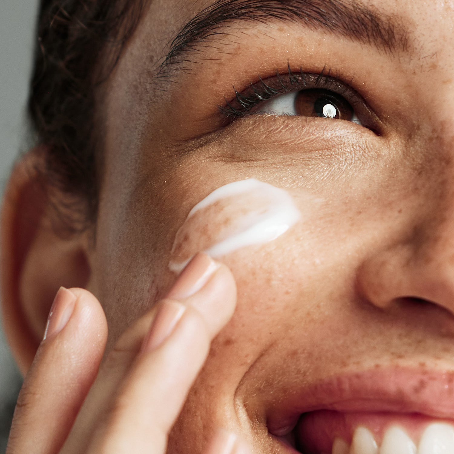 An image of a person smiling while applying a white moisturizer to their cheek with their fingertips